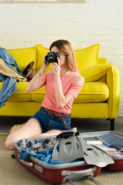 Young girl taking photo on camera while packing travel bag at home — Stock Photo