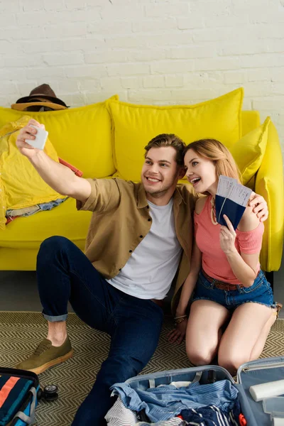 Pareja joven tomando selfie en el teléfono inteligente mientras sostiene pasaportes y billetes de avión - foto de stock