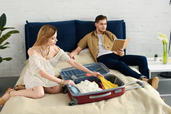 Mujer embarazada embalaje bolsa de viaje para vacaciones mientras que el marido lee libro en el dormitorio - foto de stock