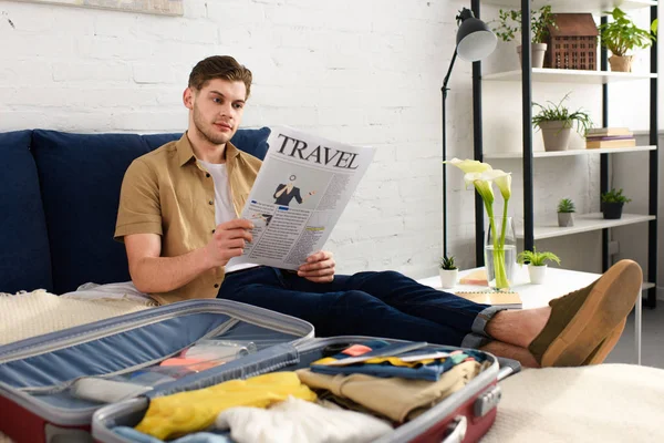 Молодой человек читает туристическую газету, упаковывая чемодан на кровати — стоковое фото