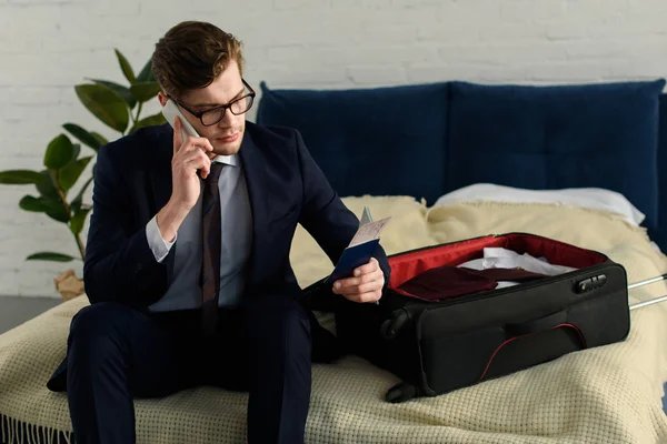 Красивый бизнесмен, держащий билет на самолет с паспортом и разговаривающий по смартфону, сидя на плохом чемодане — стоковое фото