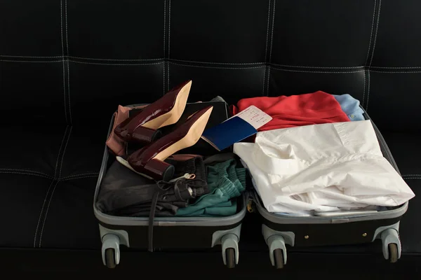Bolsa de viaje abierta con pasaporte, ropa y tacones en el sofá - foto de stock