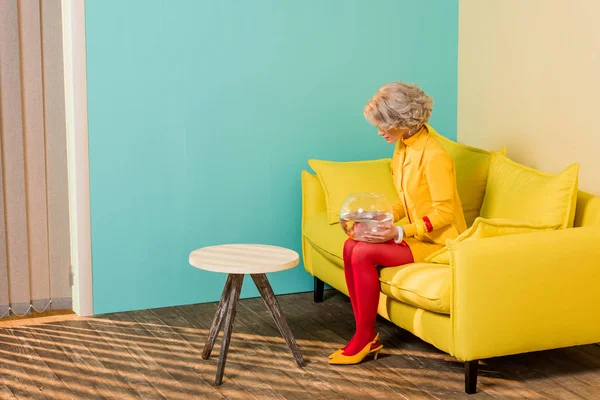 Femme dans des vêtements rétro lumineux avec du poisson doré dans l'aquarium reposant sur un canapé dans un appartement coloré, concept de maison de poupée — Photo de stock