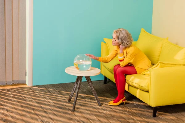 Mulher em roupas retro brilhante olhando para peixes de aquário enquanto descansa no sofá no apartamento colorido, conceito de casa de boneca — Fotografia de Stock