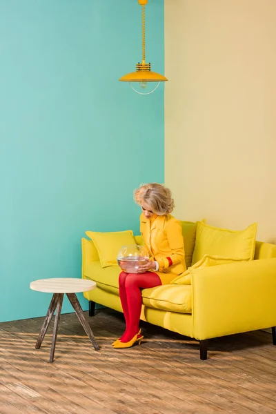 Femme dans des vêtements rétro lumineux avec du poisson doré dans l'aquarium reposant sur un canapé dans un appartement coloré, concept de maison de poupée — Photo de stock