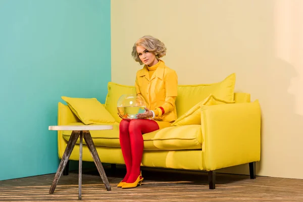 Femme en vêtements rétro lumineux avec poisson doré dans l'aquarium sur canapé à l'appartement coloré, concept de maison de poupée — Photo de stock