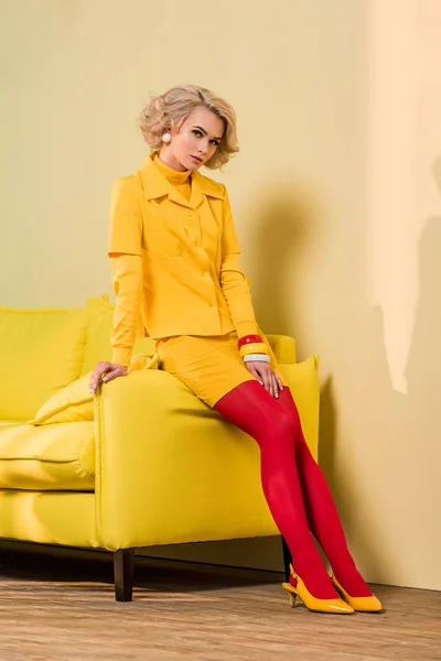 Jeune femme réfléchie dans des vêtements rétro assis sur un canapé jaune, concept de maison de poupée — Photo de stock
