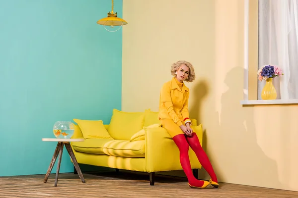 Mujer joven pensativa en ropa retro sentada en un sofá amarillo en un apartamento colorido, concepto de casa de muñecas - foto de stock