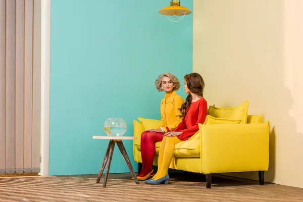 Mujeres de estilo retro sentadas en un sofá amarillo con peces de acuario en la mesa de café en el apartamento colorido, concepto de casa de muñecas - foto de stock