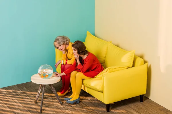 Mujeres de estilo retro sentadas en un sofá amarillo con peces de acuario en la mesa de café en el apartamento colorido, concepto de casa de muñecas - foto de stock