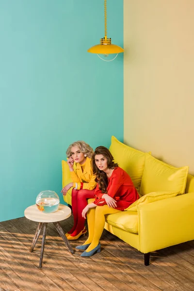Жінки в стилі ретро, що сидять на жовтому дивані з акваріумною рибою на кавовому столі за барвистою квартирою, концепція лялькового будинку — стокове фото