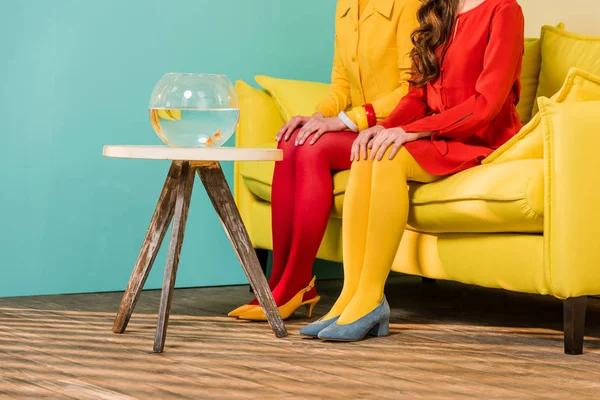 Vista parcial de mujeres de estilo retro sentadas en un sofá amarillo con peces de acuario en la mesa de café en un apartamento colorido, concepto de casa de muñecas - foto de stock