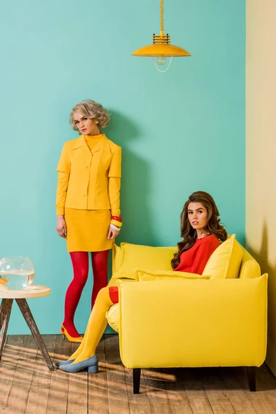 Mujeres jóvenes de estilo retro en apartamento colorido con sofá amarillo y peces de acuario, concepto de casa de muñecas - foto de stock