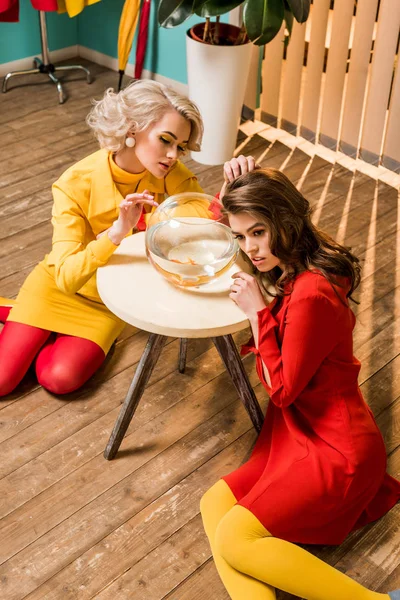 Jovem retro estilo mulheres sentadas no chão perto de peixes dourados em aquário na mesa de café, conceito de casa de boneca — Fotografia de Stock