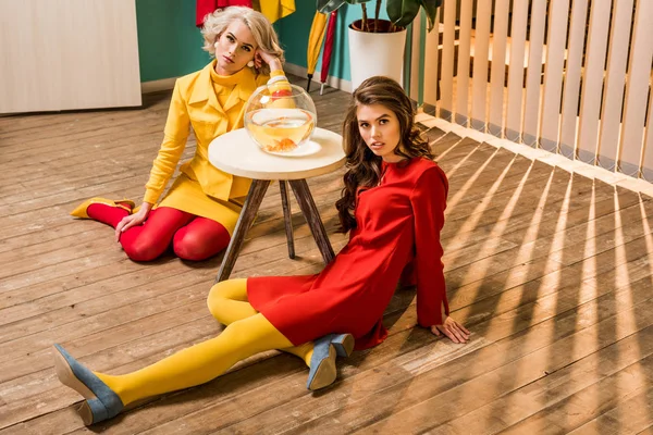 Giovani donne in stile retrò sedute sul pavimento vicino a pesci dorati in acquario sul tavolino, concetto di casa bambola — Foto stock