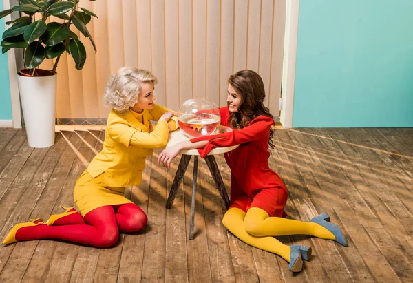 Smiling retro styled women sitting on floor near golden fish in aquarium on coffee table, doll house concept — Stock Photo