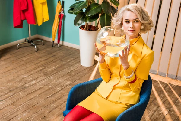 Porträt einer Frau in Retro-Kleidung mit goldenen Fischen im Aquarium auf Stuhl in buntem Appartement sitzend, Puppenhauskonzept — Stockfoto