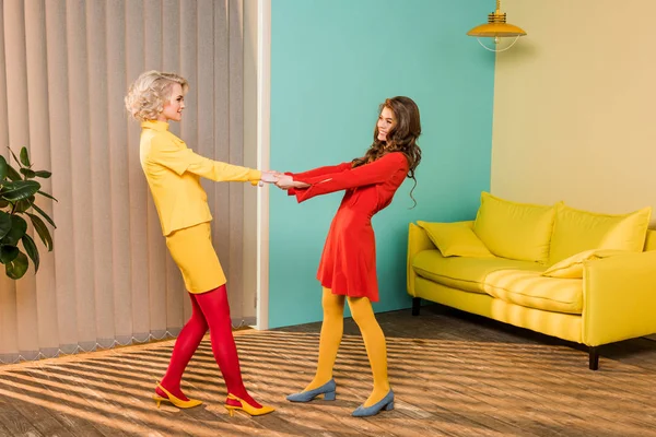 Vista lateral de mujeres bastante sonrientes en ropa retro cogidas de la mano en el apartamento colorido, concepto de casa de muñecas - foto de stock