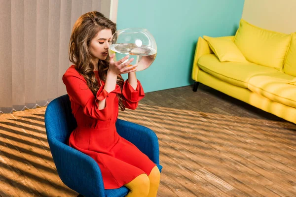 Mujer joven en ropa retro con peces de oro en el acuario sentado en la silla en el apartamento de colores, concepto de casa de muñecas - foto de stock