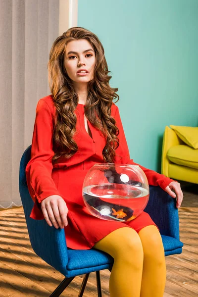 Mujer bonita en ropa retro con peces de oro en el acuario sentado en la silla en el apartamento de colores, concepto de casa de muñecas - foto de stock