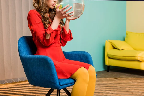 Partial view of woman in retro clothing with golden fish in aquarium sitting on chair at colorful apartment, doll house concept — Stock Photo