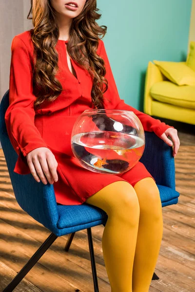 Частичный вид женщины в ретро-одежде с золотой рыбой в аквариуме, сидящей на стуле в красочной квартире, кукольный дом концепции — стоковое фото