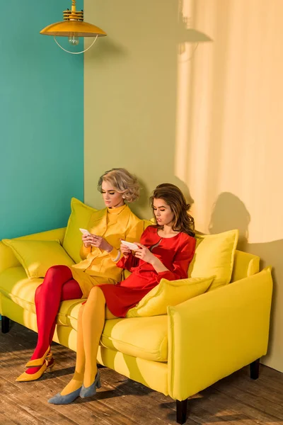 Mujeres de estilo retro utilizando teléfonos inteligentes mientras descansa en el sofá amarillo, concepto de casa de muñecas - foto de stock