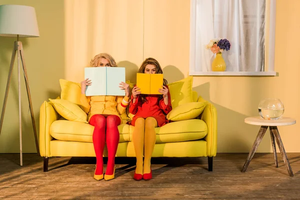 Obscured view of women in retro clothing with books sitting on yellow sofa at bright apartment, doll house concept — Stock Photo