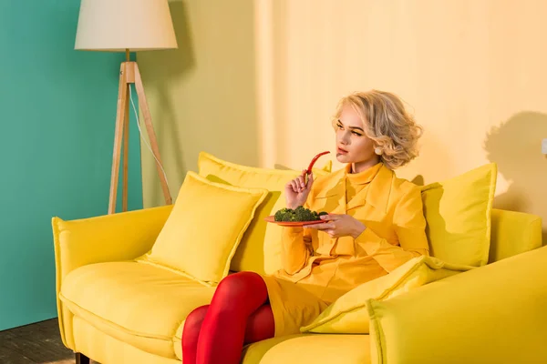 Жінка в стилі ретро з брокколі на тарілці і перець чилі в руці відпочиває на дивані в яскравій квартирі, концепція лялькового будинку — стокове фото