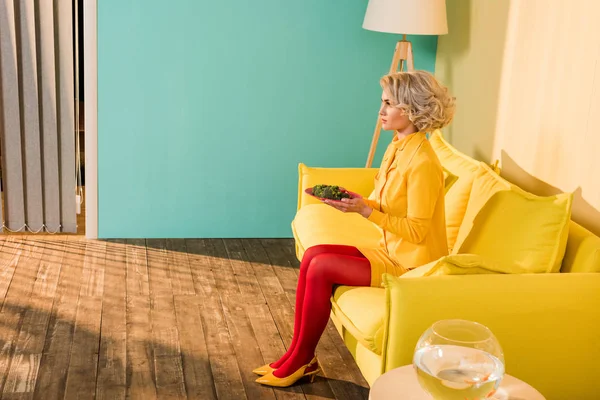 Frau im Retro-Stil mit Brokkoli auf Teller auf Sofa in heller Wohnung, Puppenhauskonzept — Stockfoto