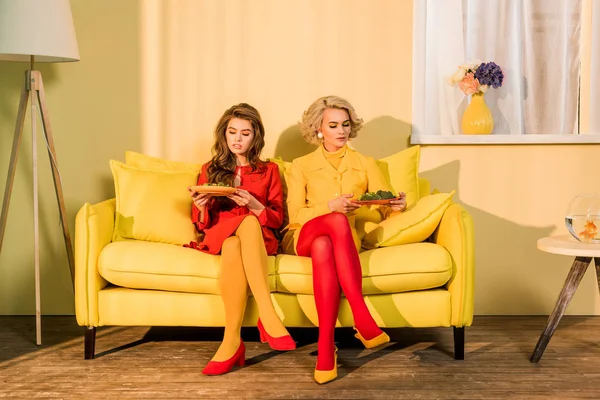 Mujeres bonitas en ropa retro con verduras en platos sentados en un sofá amarillo en una habitación luminosa, concepto de casa de muñecas - foto de stock