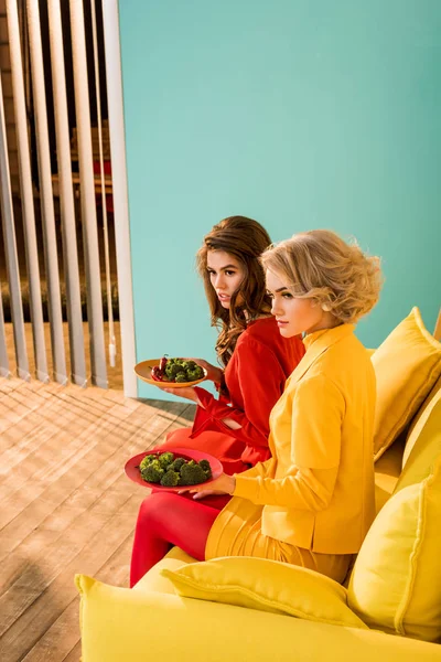 Красиві жінки в ретро одязі з овочами на тарілках, що сидять на жовтому дивані в барвистій кімнаті, концепція лялькового будинку — стокове фото