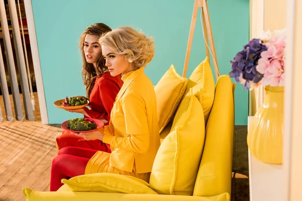 Mujeres bonitas en ropa retro con verduras en platos sentados en un sofá amarillo en una habitación colorida, concepto de casa de muñecas - foto de stock