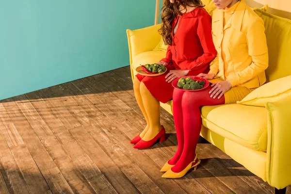 Cropped image of retro styled girls in colorful dresses sitting with plates of broccoli at home — Stock Photo