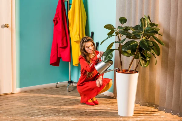 Bela mulher em vestido vermelho agachamento e regando planta com regar pode em casa — Fotografia de Stock