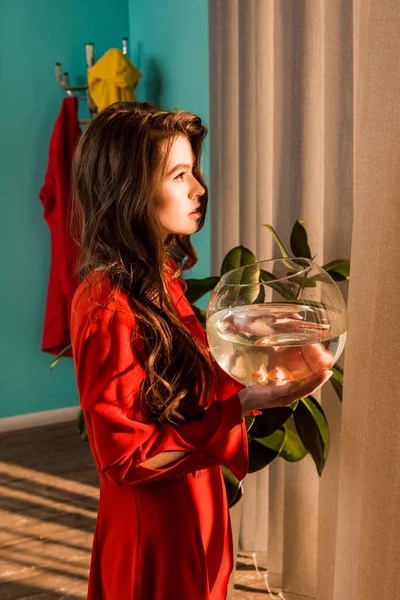 Vue latérale de la femme élégante en robe rouge tenant aquarium avec des poissons d'or et regardant loin à la maison — Photo de stock