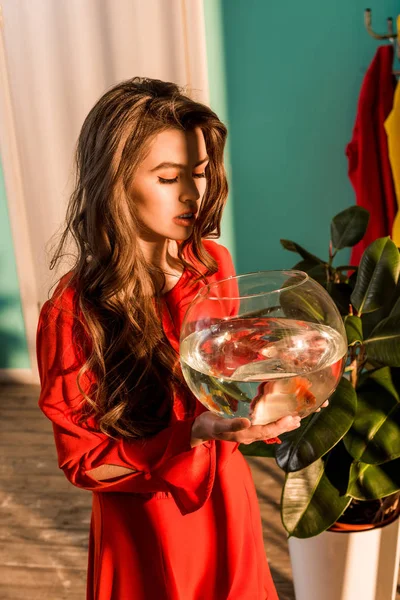 Femme à l'ancienne en robe rouge regardant l'aquarium avec des poissons d'or à la maison — Photo de stock