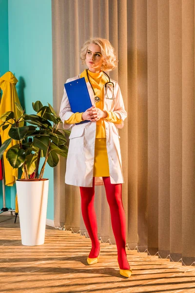 Beautiful retro styled doctor in colorful dress holding clipboard and looking away in clinic — Stock Photo