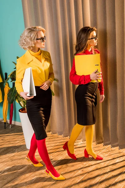 Hermosas mujeres de negocios de estilo retro en coloridas blusas caminando con carpetas en la oficina - foto de stock