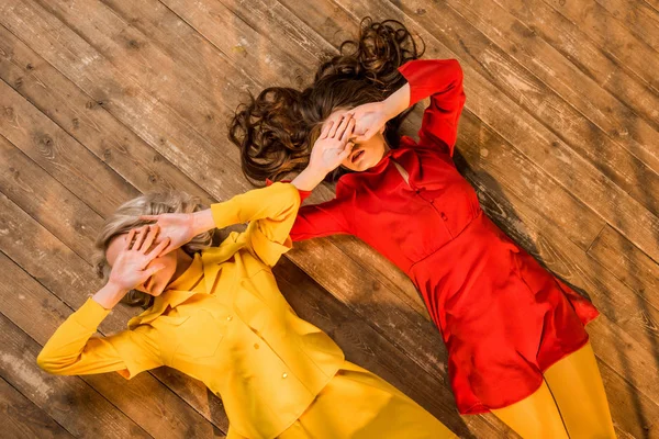 Vista superior de las niñas de estilo retro en vestidos de colores tumbados en el suelo y cubriendo los ojos con las manos en casa - foto de stock