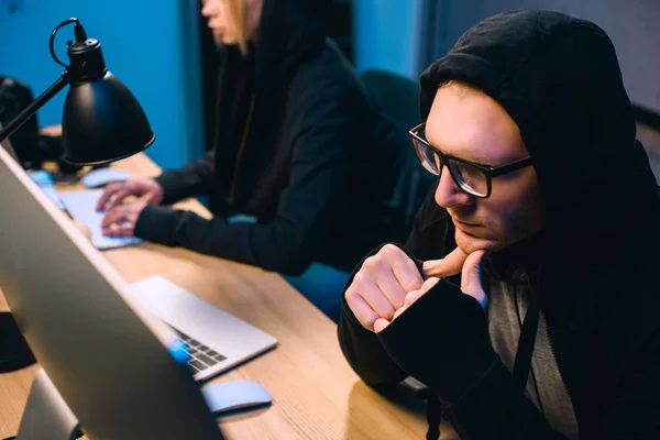 Nachdenklicher junger Hacker blickt in dunklen Raum auf Computerbildschirm — Stockfoto