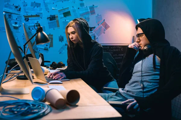 Pareja de jóvenes piratas informáticos que utilizan el ordenador portátil en el lugar de trabajo en habitación oscura - foto de stock