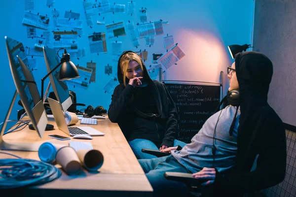 Pareja de jóvenes hackers charlando en el lugar de trabajo en habitación oscura - foto de stock