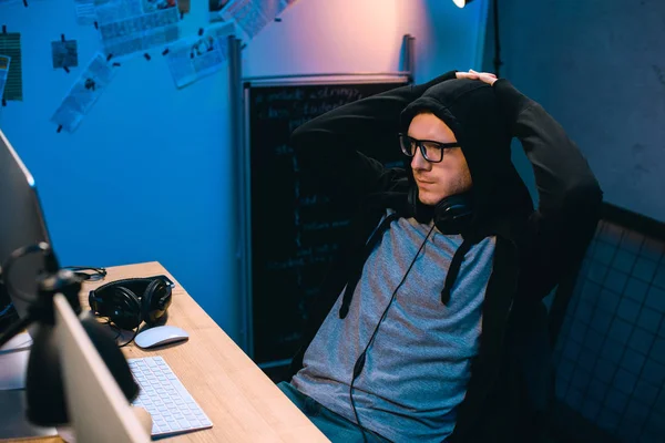 Hacker encapuchado relajarse en la silla delante de su lugar de trabajo - foto de stock