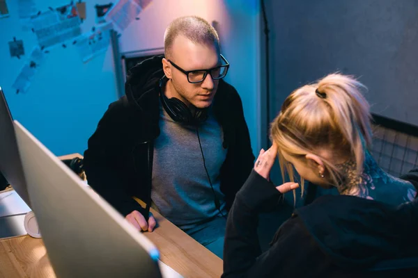 Young hacker talking with depressed accomplice at workplace in dark room — Stock Photo