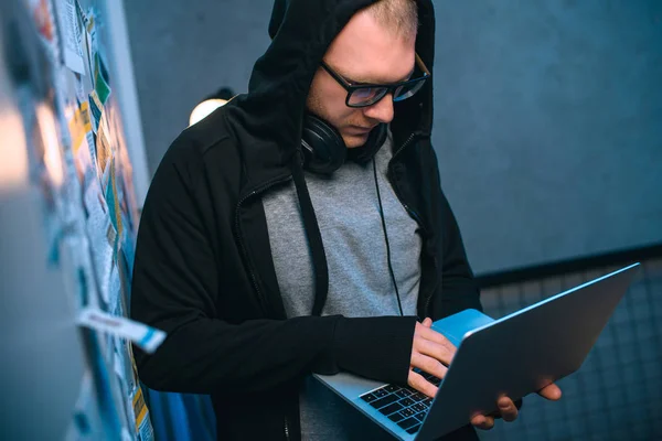 Hooded hacker developing malware with laptop in dark room — Stock Photo