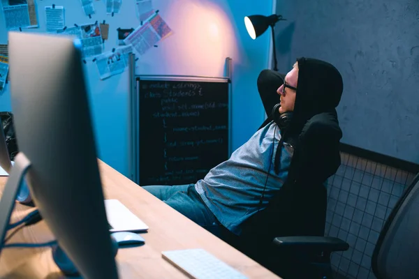 Joven hacker encapuchado relajarse en la silla delante de su lugar de trabajo - foto de stock