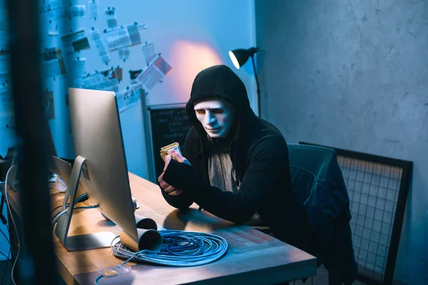 Hacker encapuchado en máscara contando dinero robado en su lugar de trabajo - foto de stock