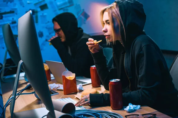 Hacker mujer que trabaja en malware con cómplice y comer comida chatarra - foto de stock
