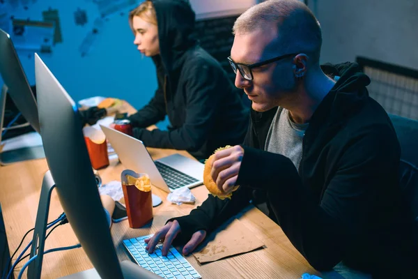 Male hacker working on malware with accomplice and eating junk food — Stock Photo